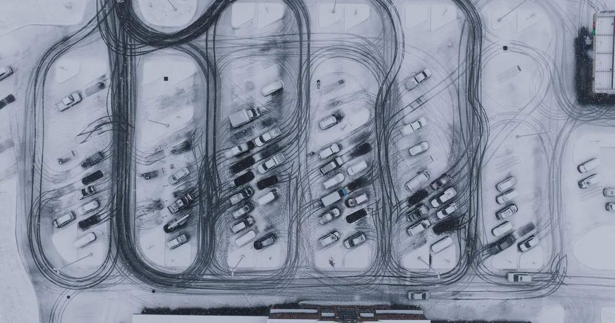Aerial view of a snowy parking lot with cars and tire tracks visible.