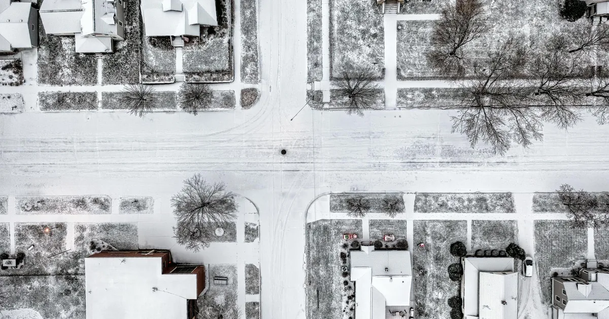 Aerial view of a snowy residential area with parked cars and tree.