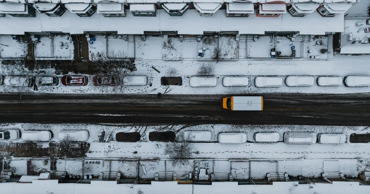 Aerial view of a snowy street with parked vehicles and residential.