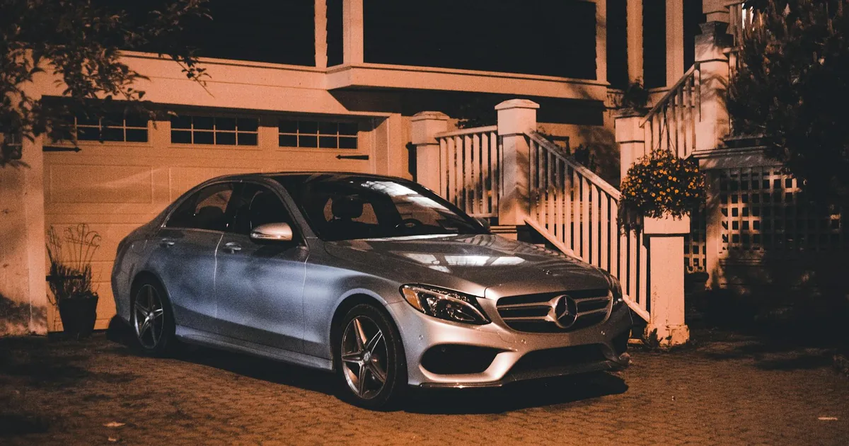 Car parked on a driveway beside a house at night with porch lights.