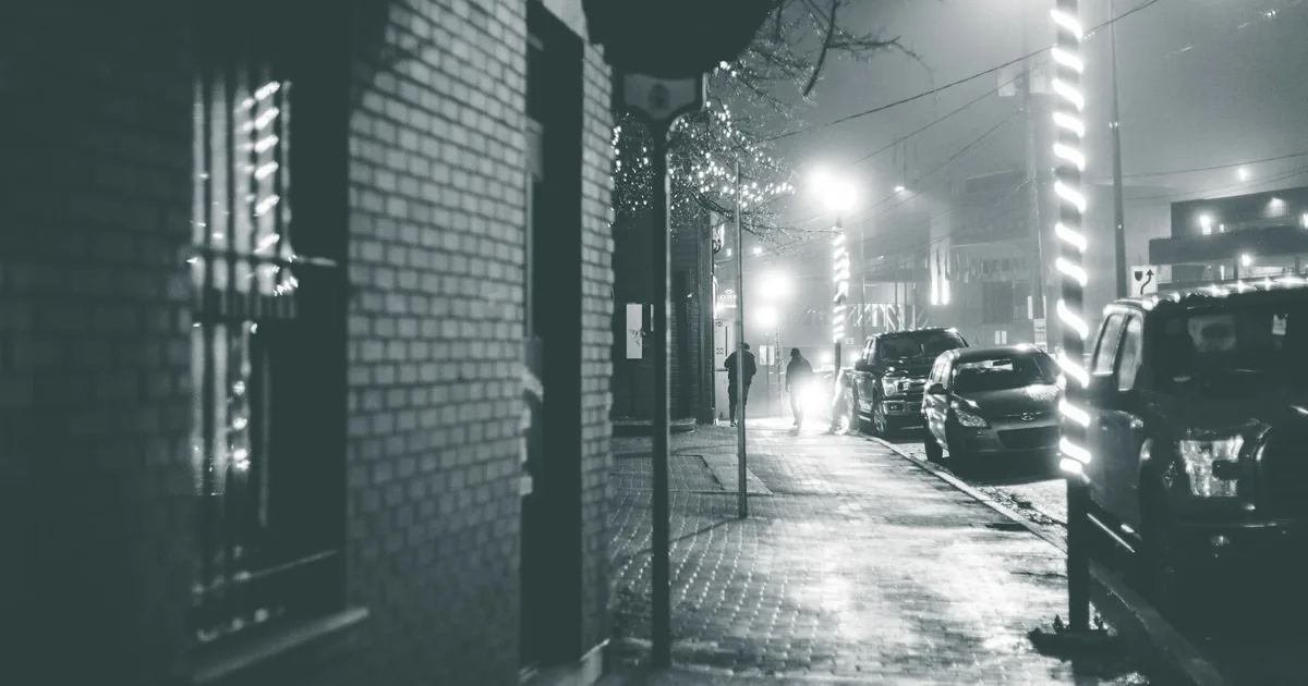 Dimly lit street with parked cars and pedestrians walking on the.