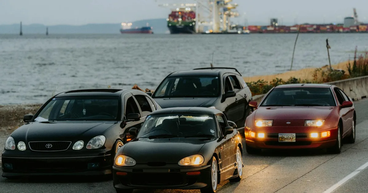 Four cars parked along a waterfront road with a shipping dock in.