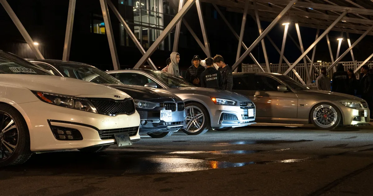 Group of parked cars under a modern structure at night with people.
