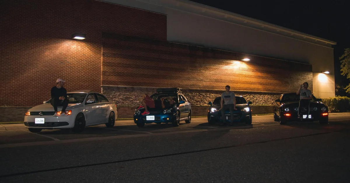 Group of people sitting on parked cars in a dimly lit parking lot.