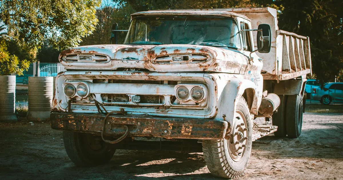 Old rusty truck parked in a gravel lot surrounded by trees.