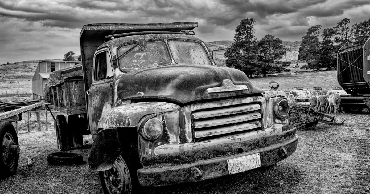 Old rusty truck parked in a rural area with trees and cloudy sky.