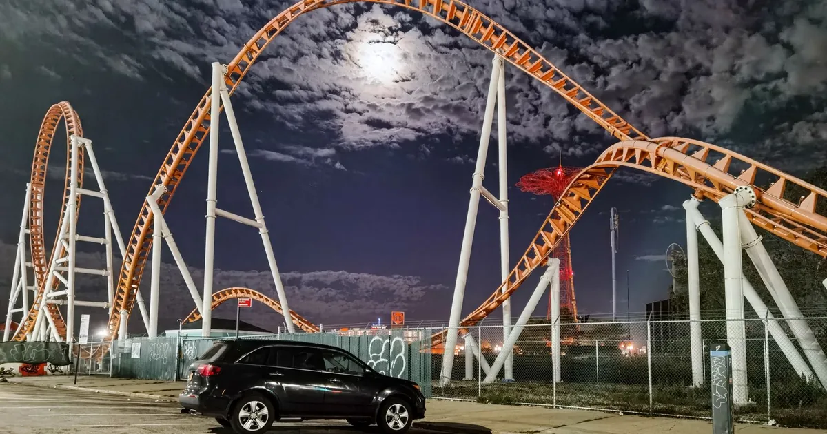 Parked car in front of a roller coaster under a night sky.