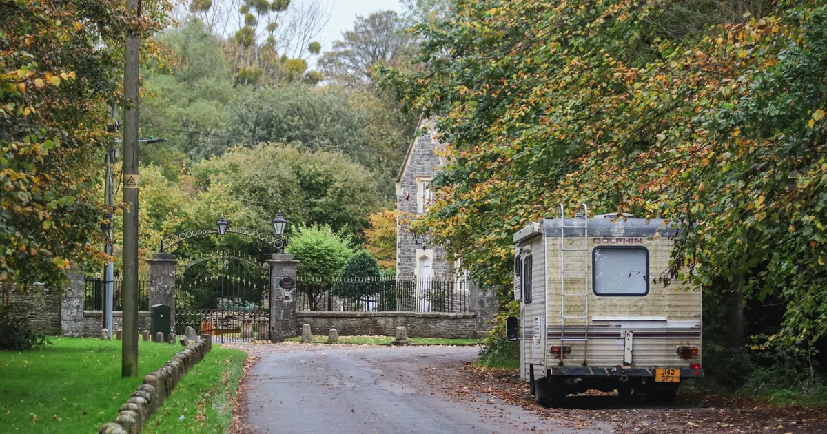 Parked vehicle beside a tree lined road near a house and gate.