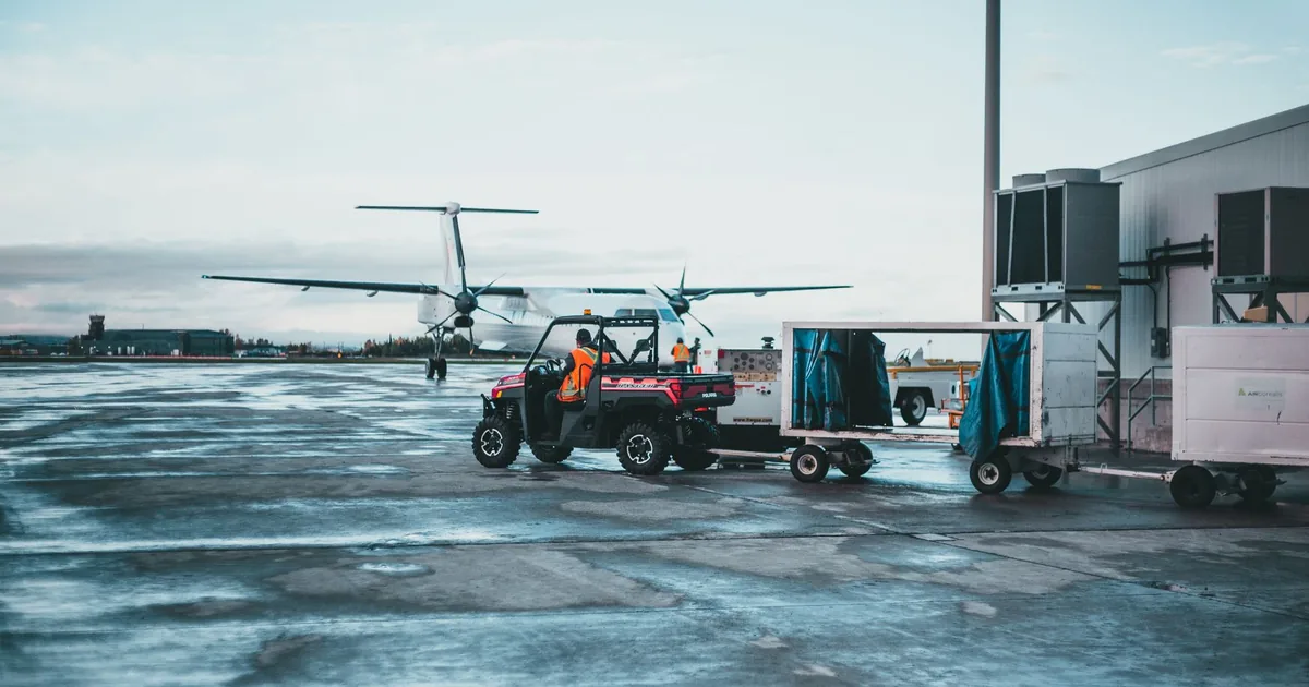Parking area at an airport with vehicles and luggage carts visible.