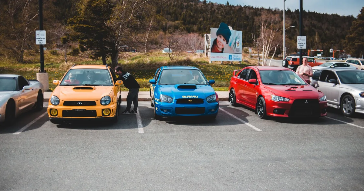 Parking lot scene with three colorful cars and people nearby.