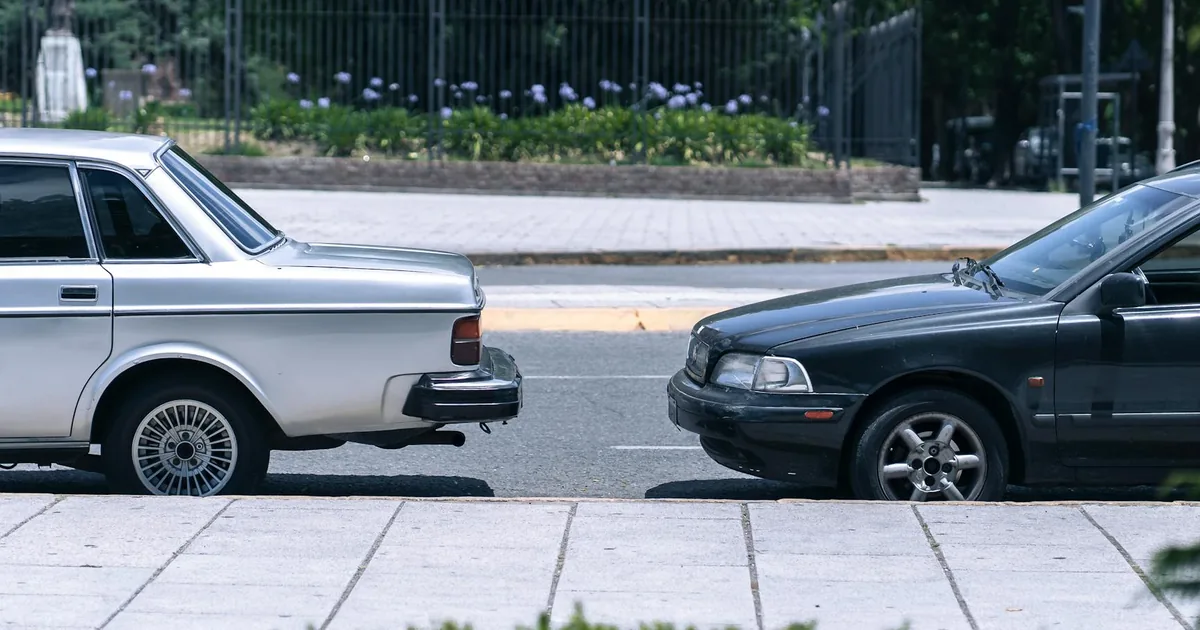 Silver car and a black car parked on a city street near greenery.