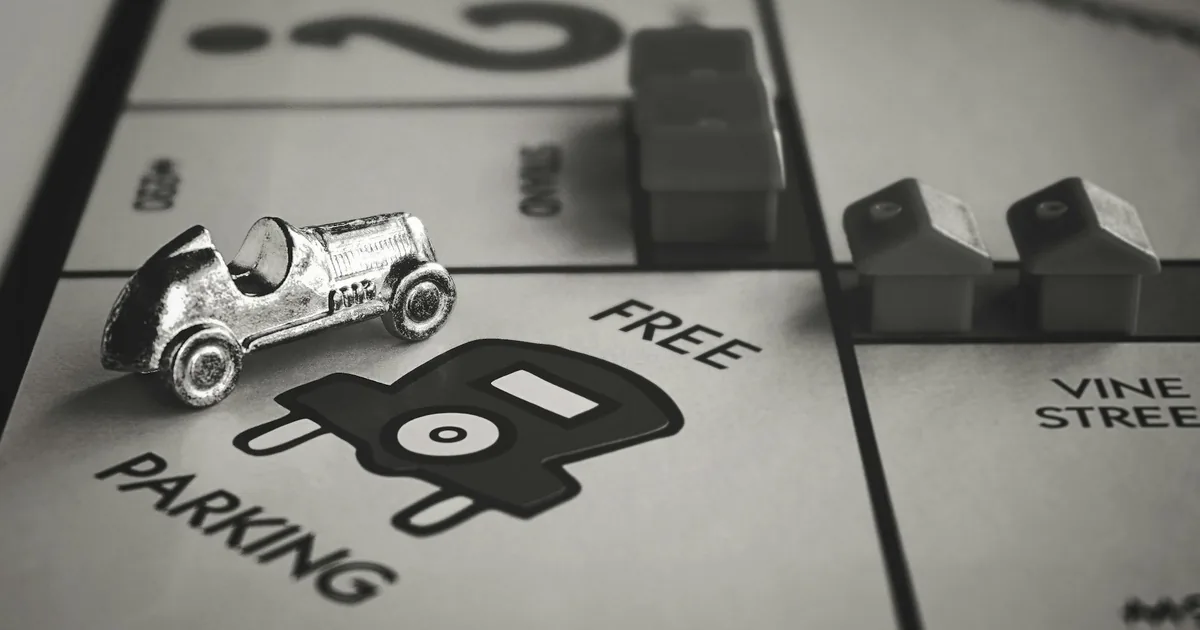 Silver car token on a monopoly board near a parking space marked.
