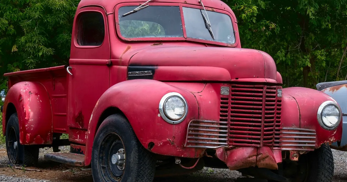 Vintage red truck parked in a gravel lot surrounded by greenery.