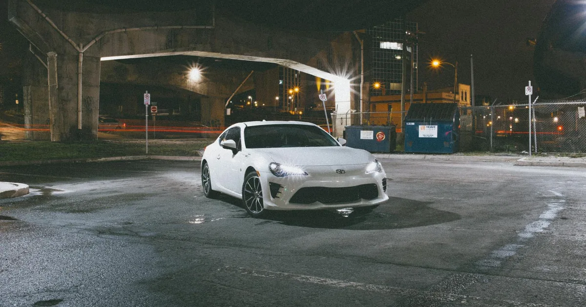 White car parked in a dimly lit urban area with nearby dumpsters.