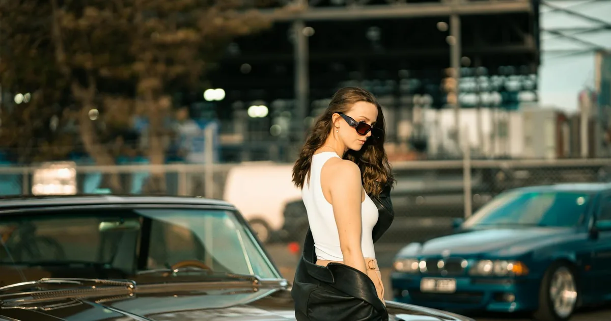 Woman in sunglasses stands beside a parked car in a busy parking lot.