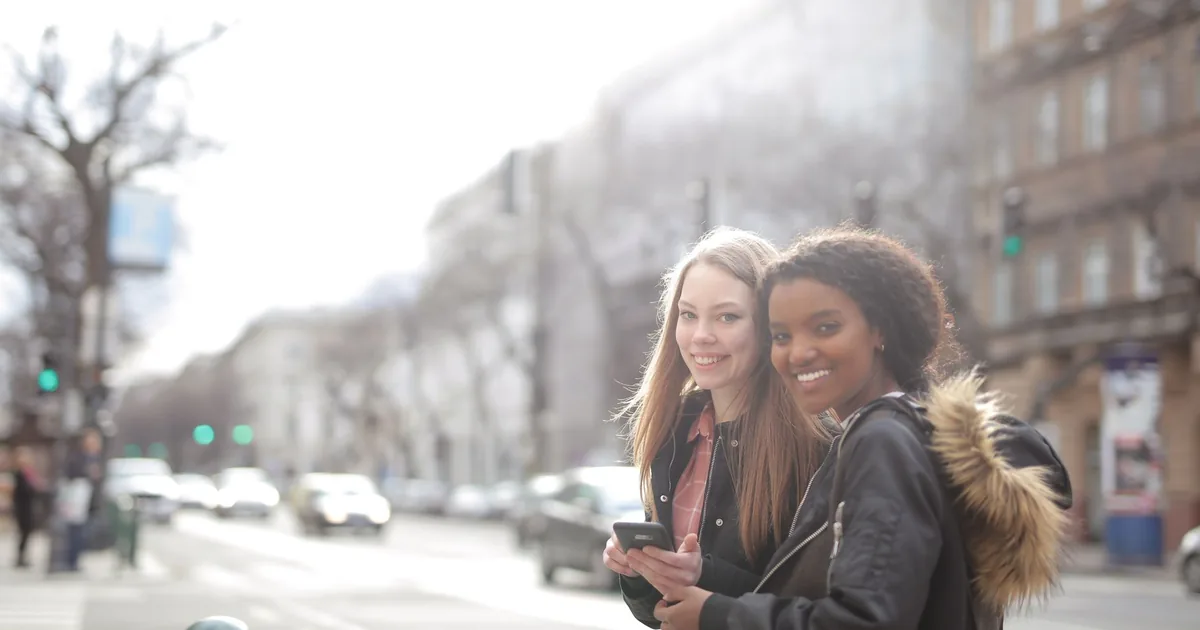 Two women smiling and holding their phones on a busy city street