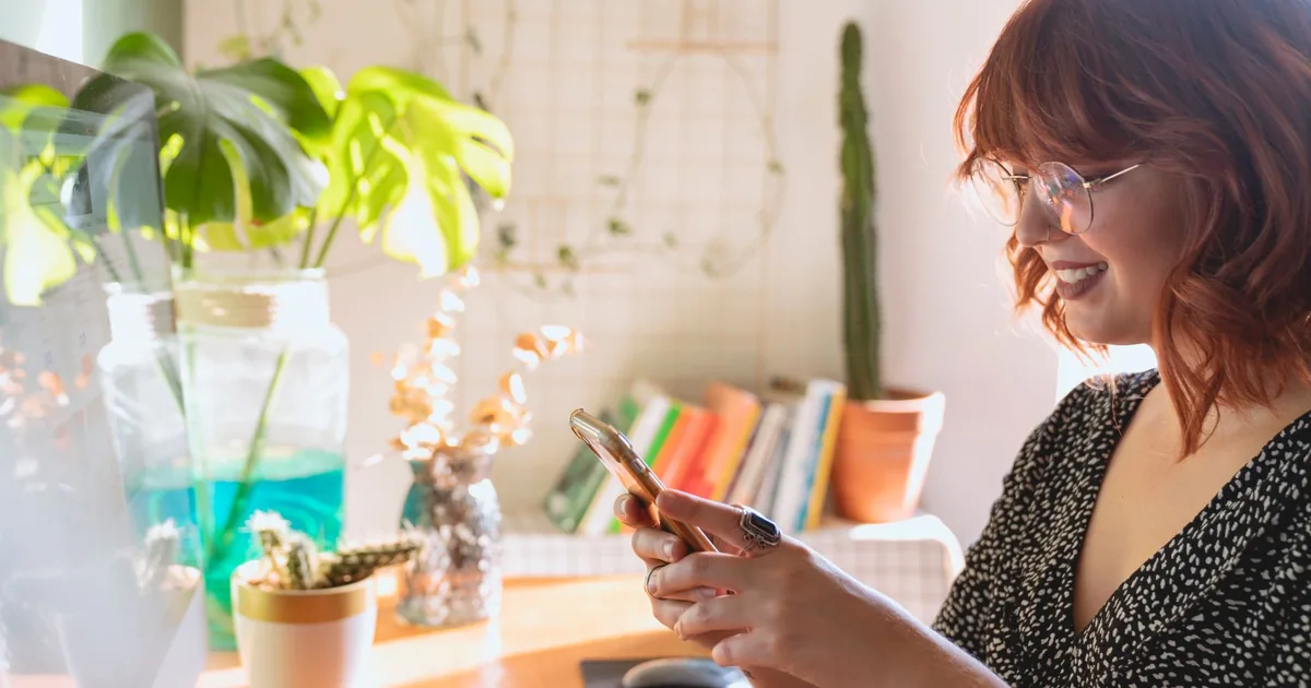 A woman smiling while using her smartphone at a desk at home